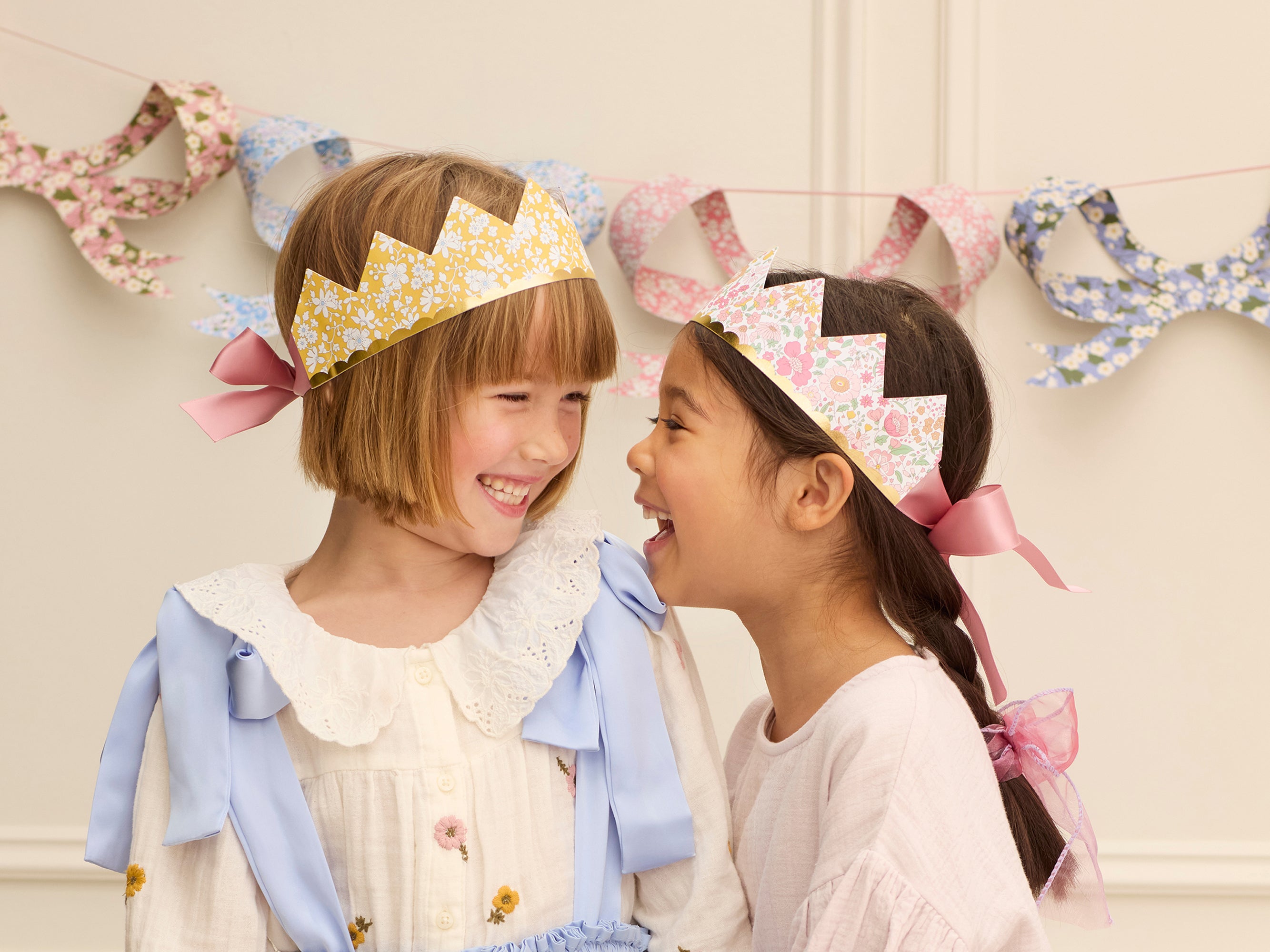 Two children wearing decorative crowns with a floral and ribbon design, standing against a light-coloured wall with decorative ribbons hanging | Meri Meri x Liberty Party Crowns (8) | Holme & Moss
